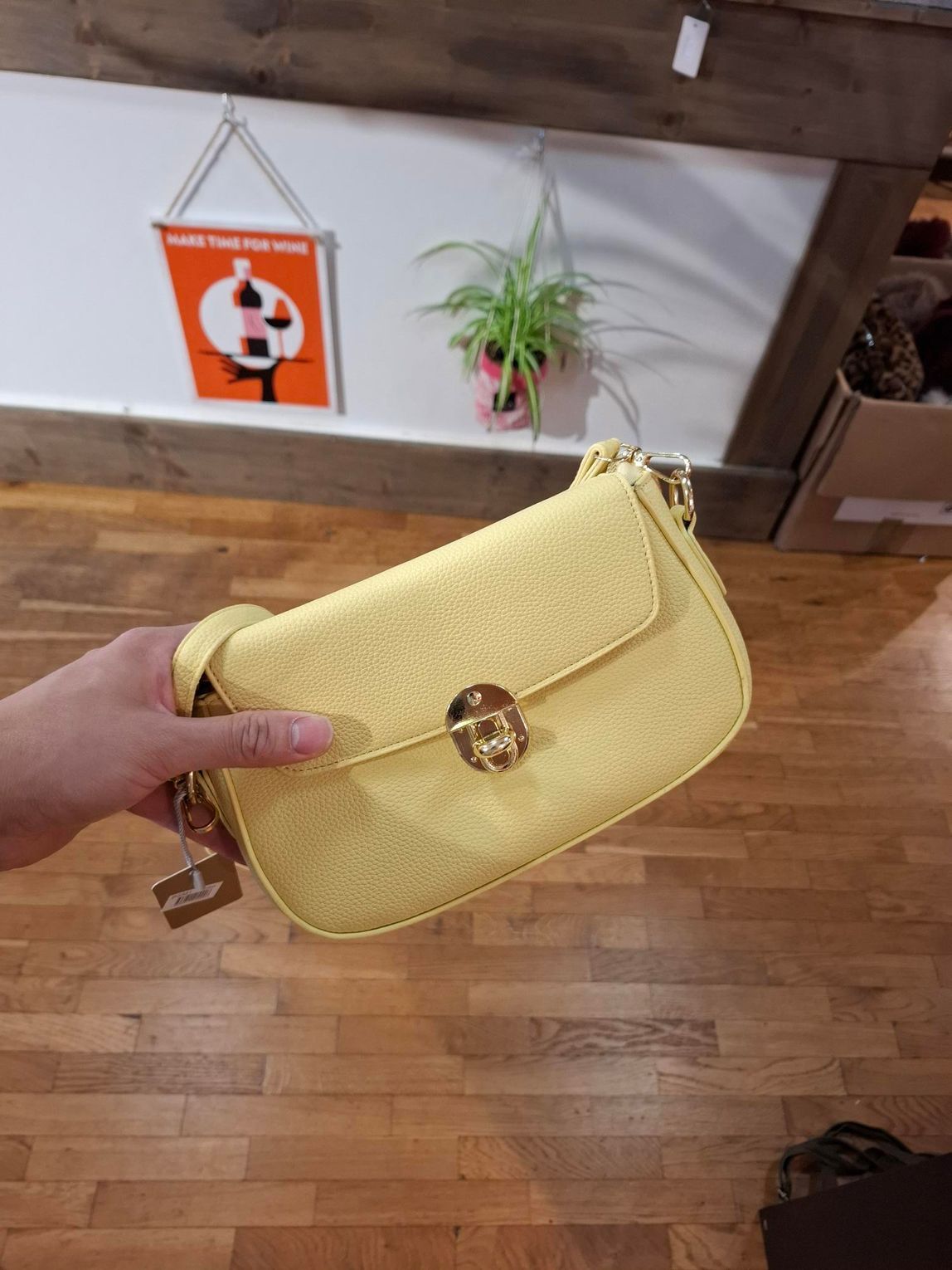 Gele handtas met gouden sluiting, die boven een houten vloer hangt, vlakbij een muurbord en een hangende plant.
