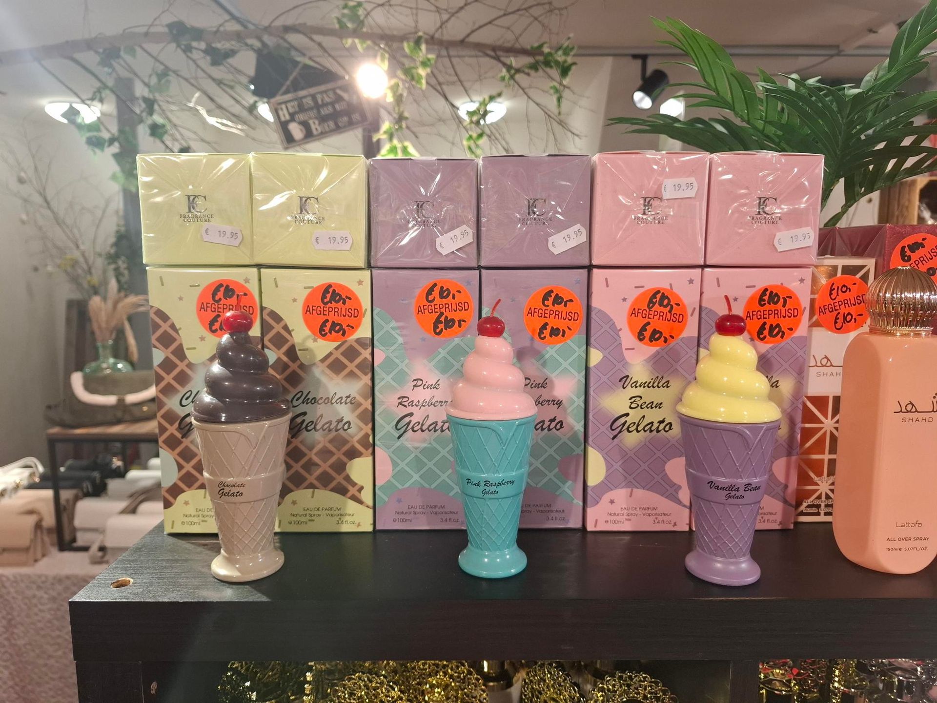 Een presentatie van pastelkleurige, ijsvormige kaarsen in wafelvormpjes op een zwarte plank, met daarachter kleurrijke verpakkingen.