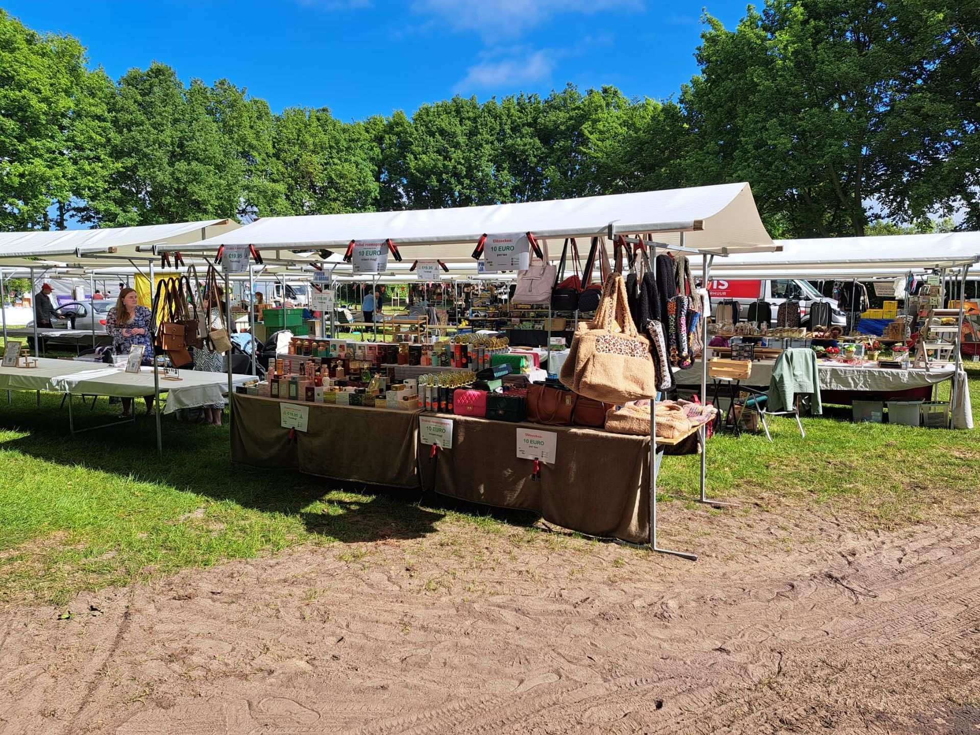 Marktstalletjes met witte luifels staan ​​opgesteld op een grasveld met bomen op de achtergrond onder een blauwe hemel.