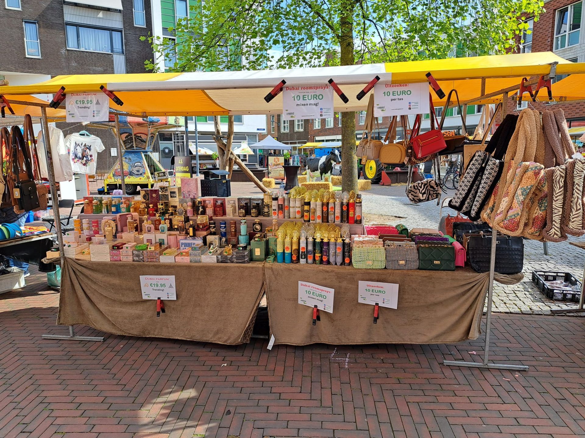 Op een marktkraam staan ​​diverse tassen, accessoires en flessen uitgestald op een tafel onder een geel-wit gestreept afdak.