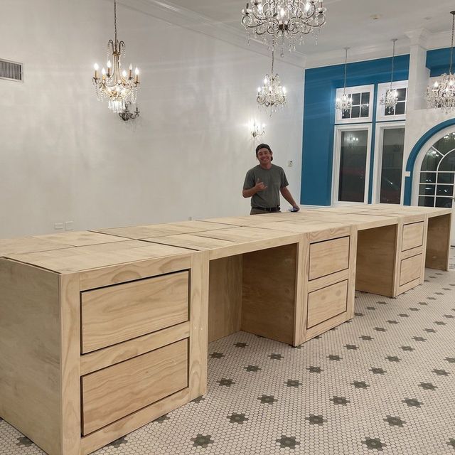 Man stands behind a long wooden desk with drawers in a room with chandeliers and teal accents.