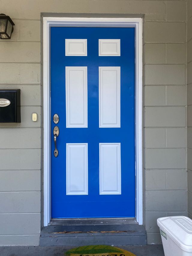 Blue front door with white trim, six panels, and silver hardware. Gray siding and a welcome mat.