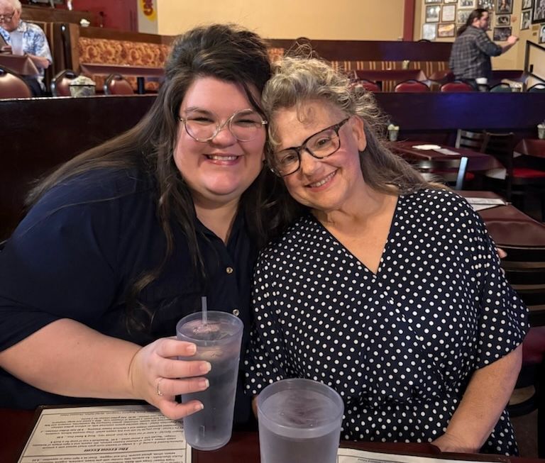 Two women smiling, hugging at a restaurant table. One holds a drink. Dark blue and polka dot tops.