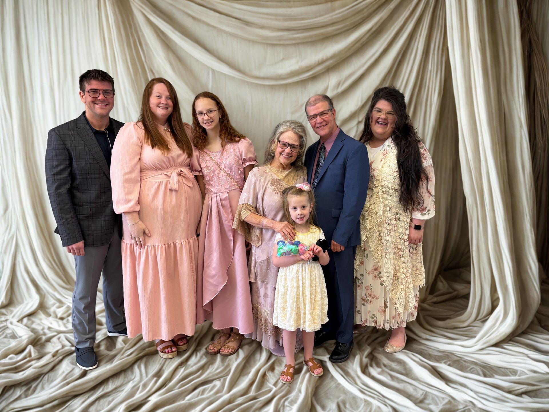 A group of seven people smiling in front of a draped, off-white fabric backdrop, posing for a portrait.