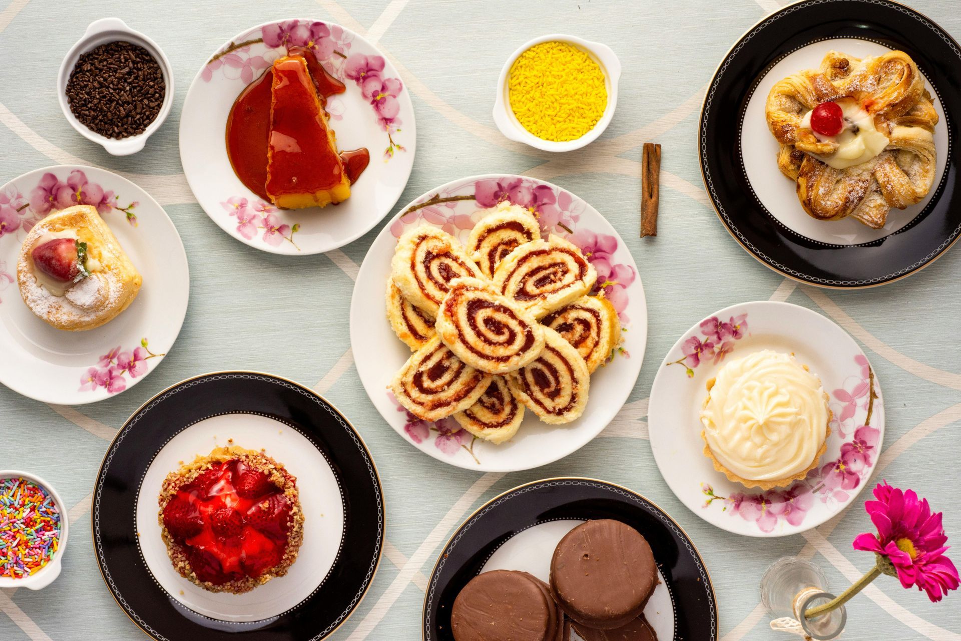 Assortment of colorful pastries on floral and black-rimmed plates, with sprinkles and cinnamon stick.