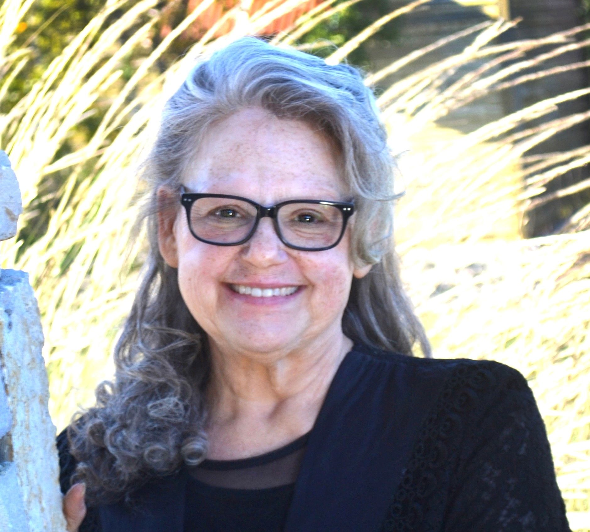 Smiling woman with glasses, gray curly hair, black top, stands outside, blurred greenery background.