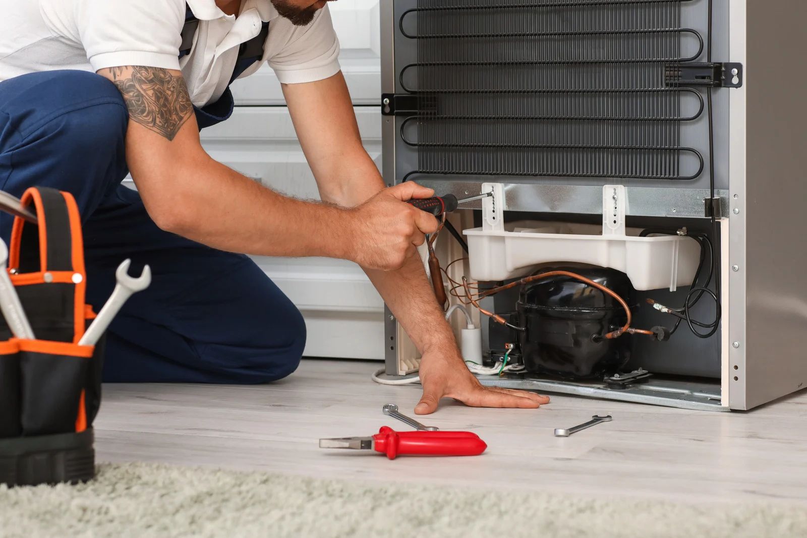 A repairman kneeling, fixing a refrigerator's compressor unit; tools sit nearby on a rug.