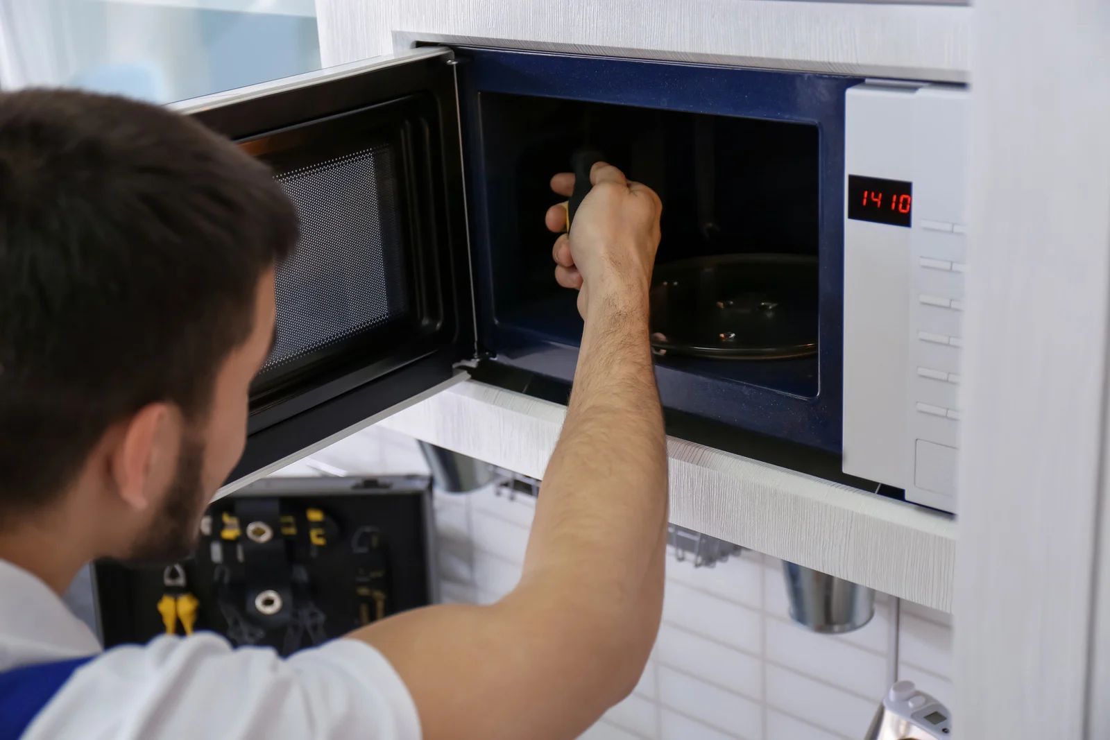 Man repairing a microwave in a kitchen; he is reaching inside with a tool.