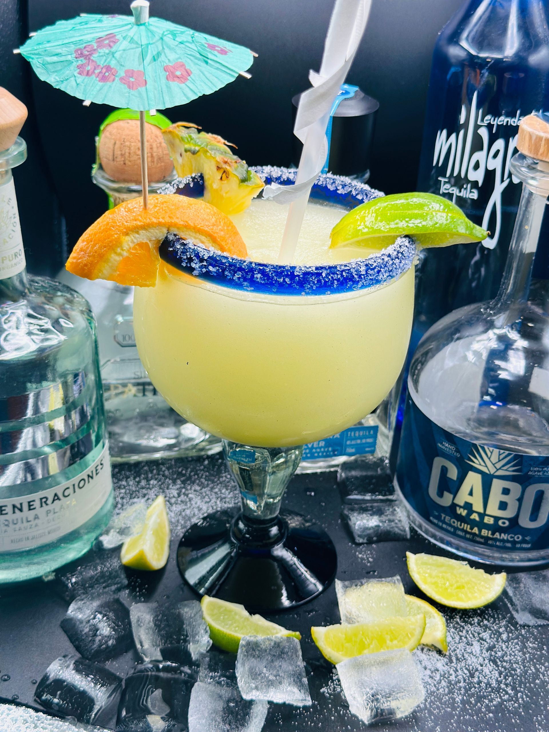 Margarita cocktail in a glass with a blue rim, garnished with fruit and an umbrella, surrounded by tequila bottles and ice.