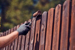 Person wearing a black glove uses a paint roller to stain a wooden fence outdoors.