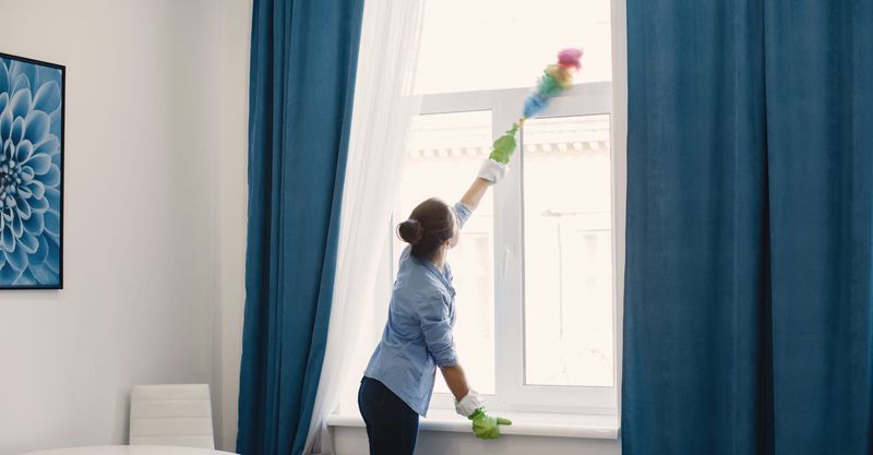 Woman dusting a window with a colorful duster in a room with blue curtains.