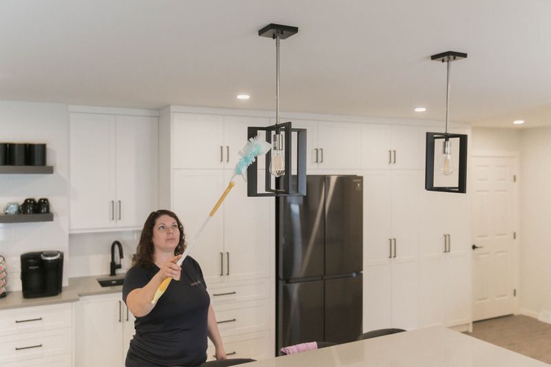 Woman dusting a black chandelier in a white kitchen with a long-handled duster.