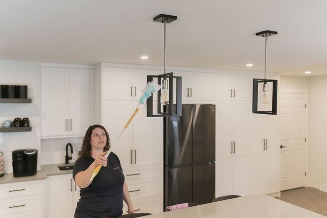 Woman dusting a black chandelier in a white kitchen with a long-handled duster.