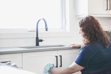 Woman wiping a white kitchen cabinet with a cloth next to a sink.