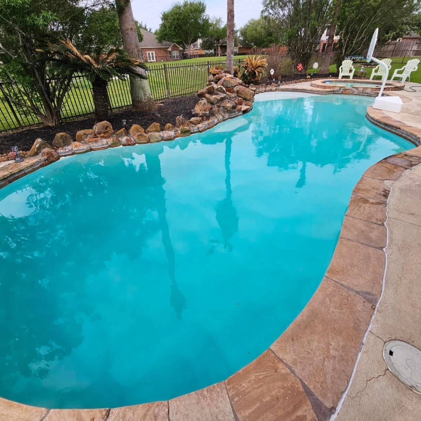 Curved backyard swimming pool with turquoise water, stone border, and patio chairs beside a grassy yard.