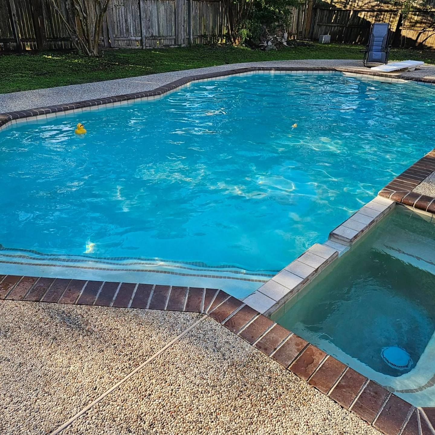 Backyard swimming pool with bright blue water and a small attached spa, bordered by brick and gravel.