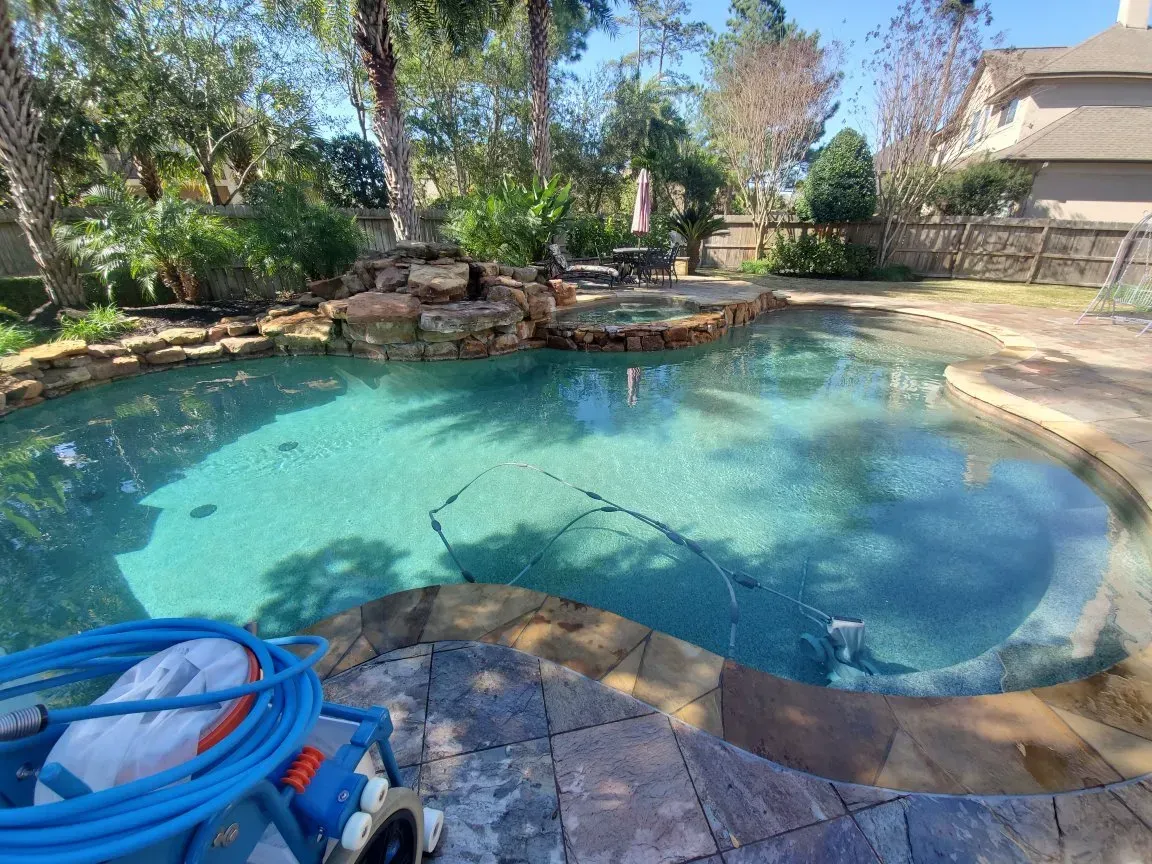 Backyard swimming pool with rock waterfall, trees, and a blue pool cleaner on the deck