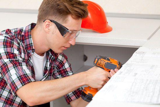 A man in a plaid shirt is using a drill under a sink.