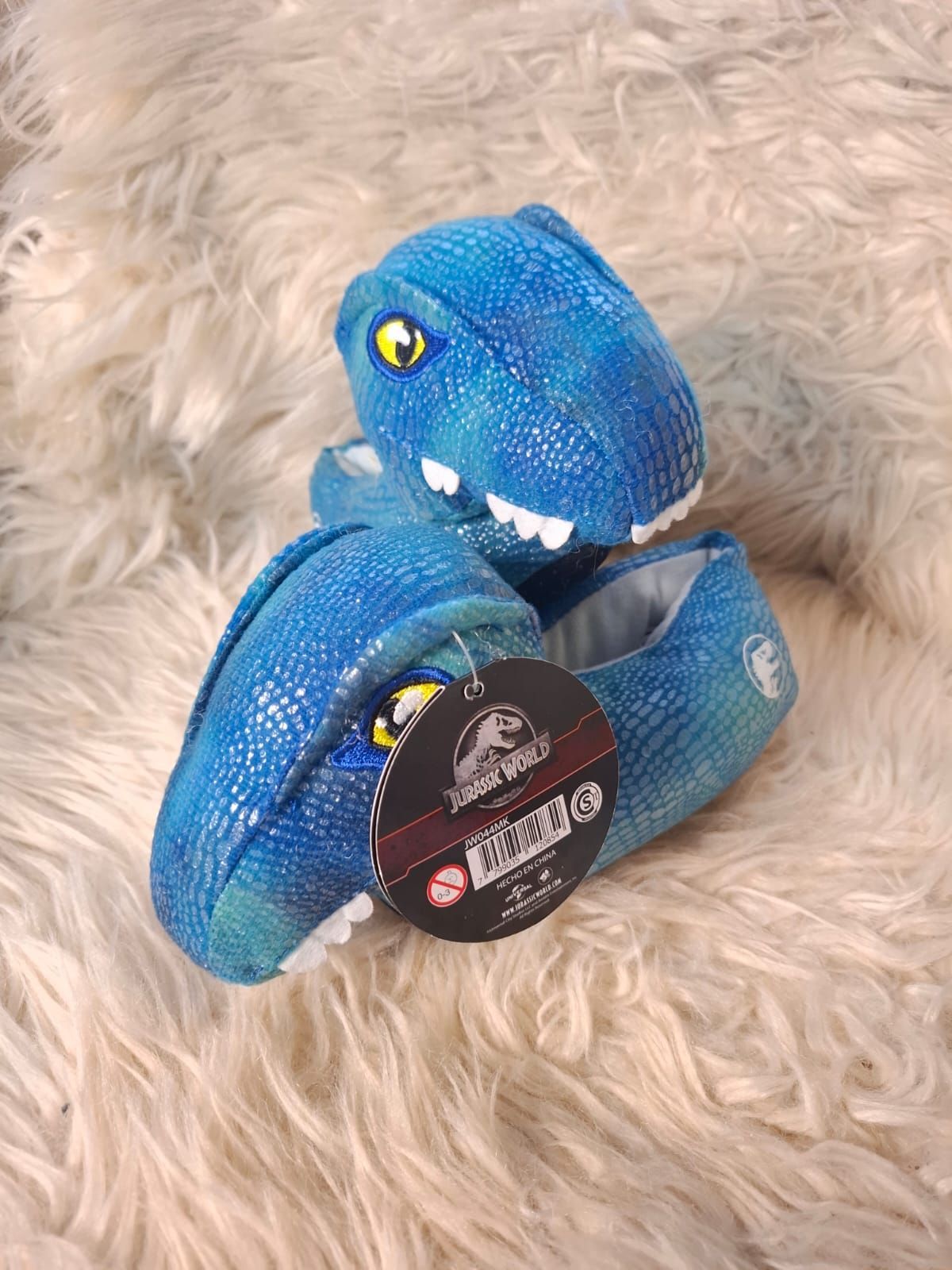 Un par de pantuflas de dinosaurio azules están colocadas encima de una superficie peluda blanca.