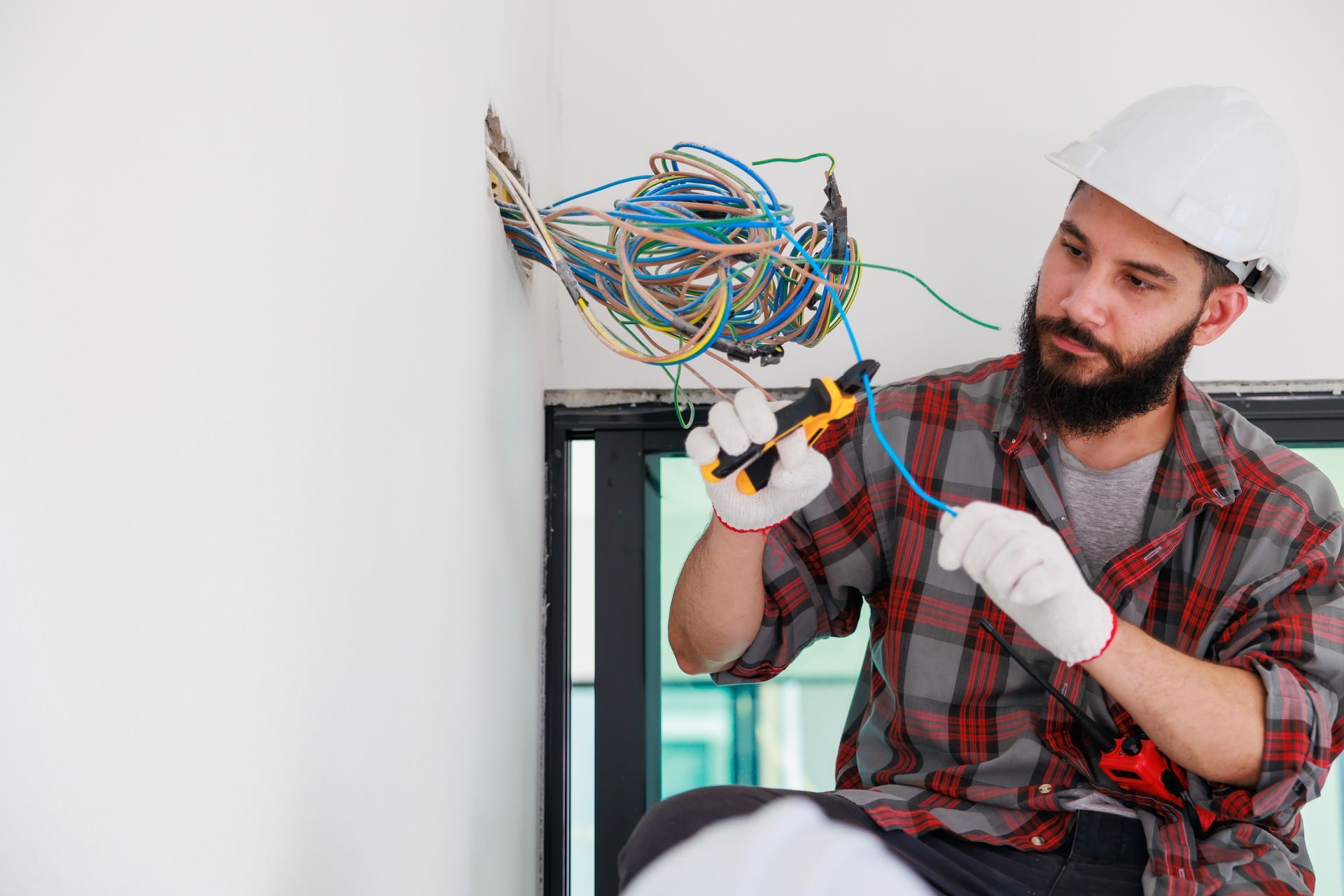 Electrician working with wires inside a wall opening. He's wearing a hard hat, gloves, and a plaid shirt.