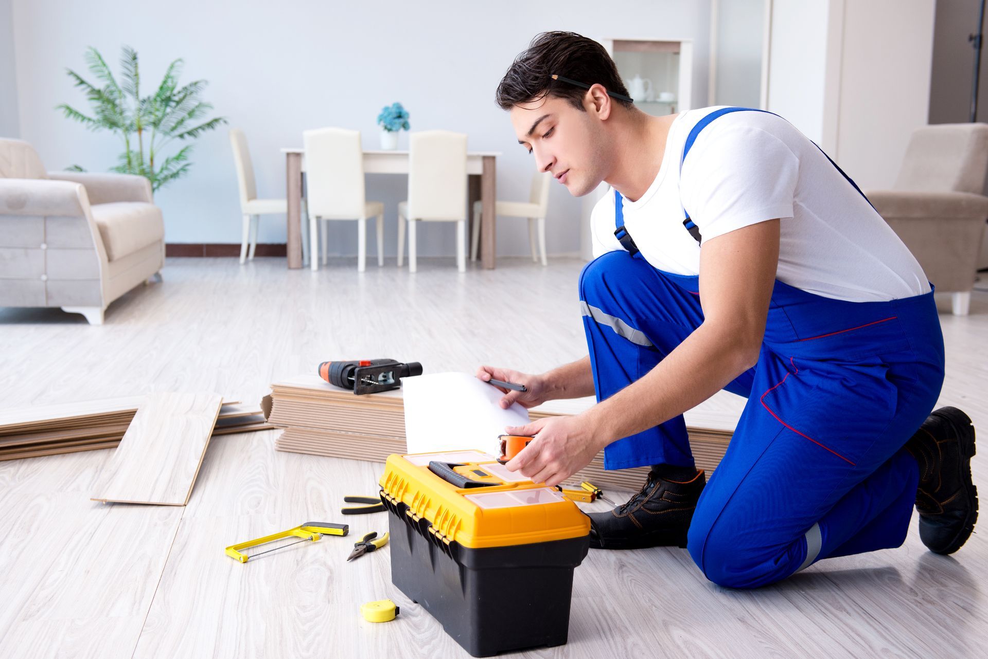A person kneels, looking at a toolbox, wood planks nearby.  They wear blue overalls in a bright living room.
