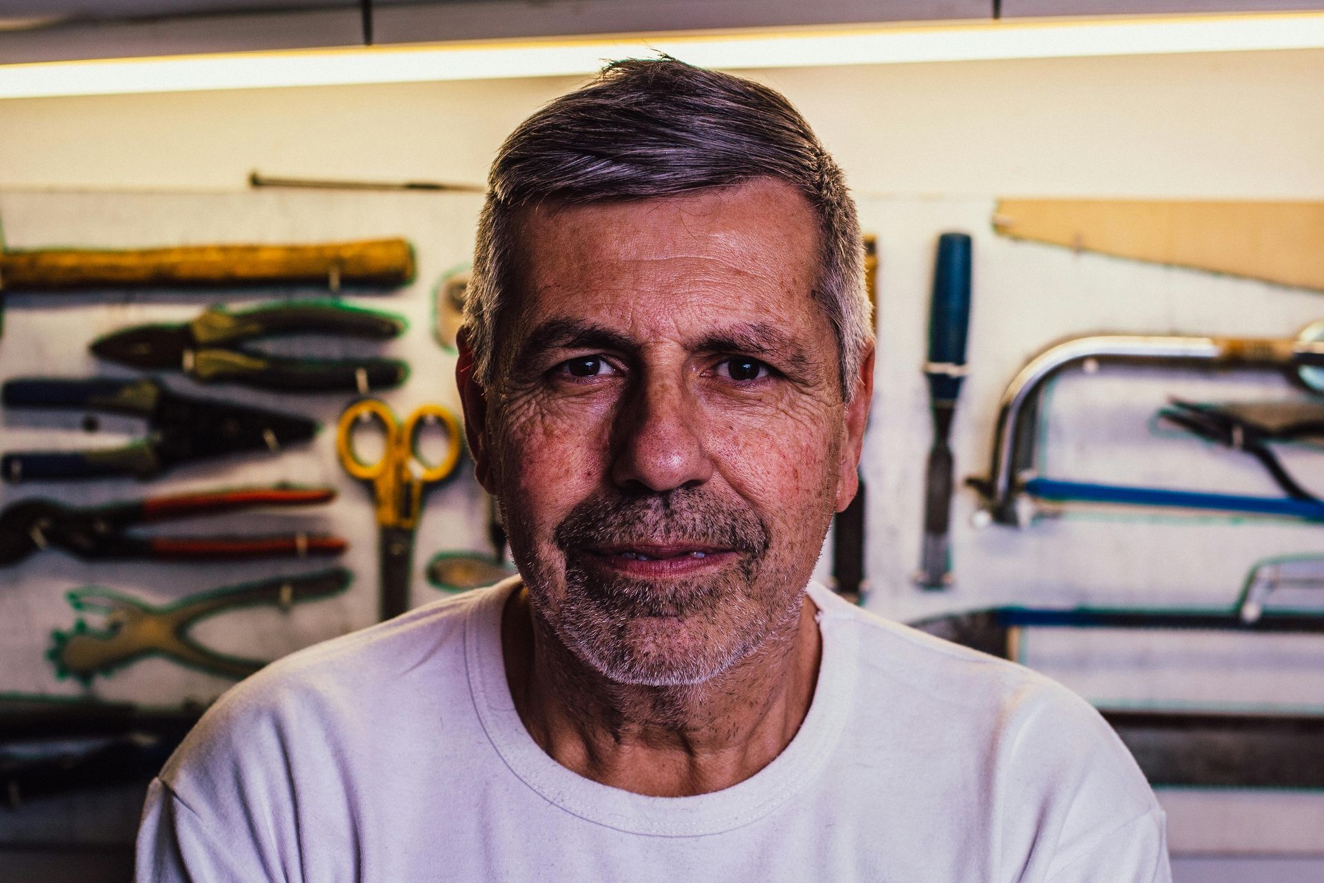 Man in white shirt smiling, workshop tools in background.