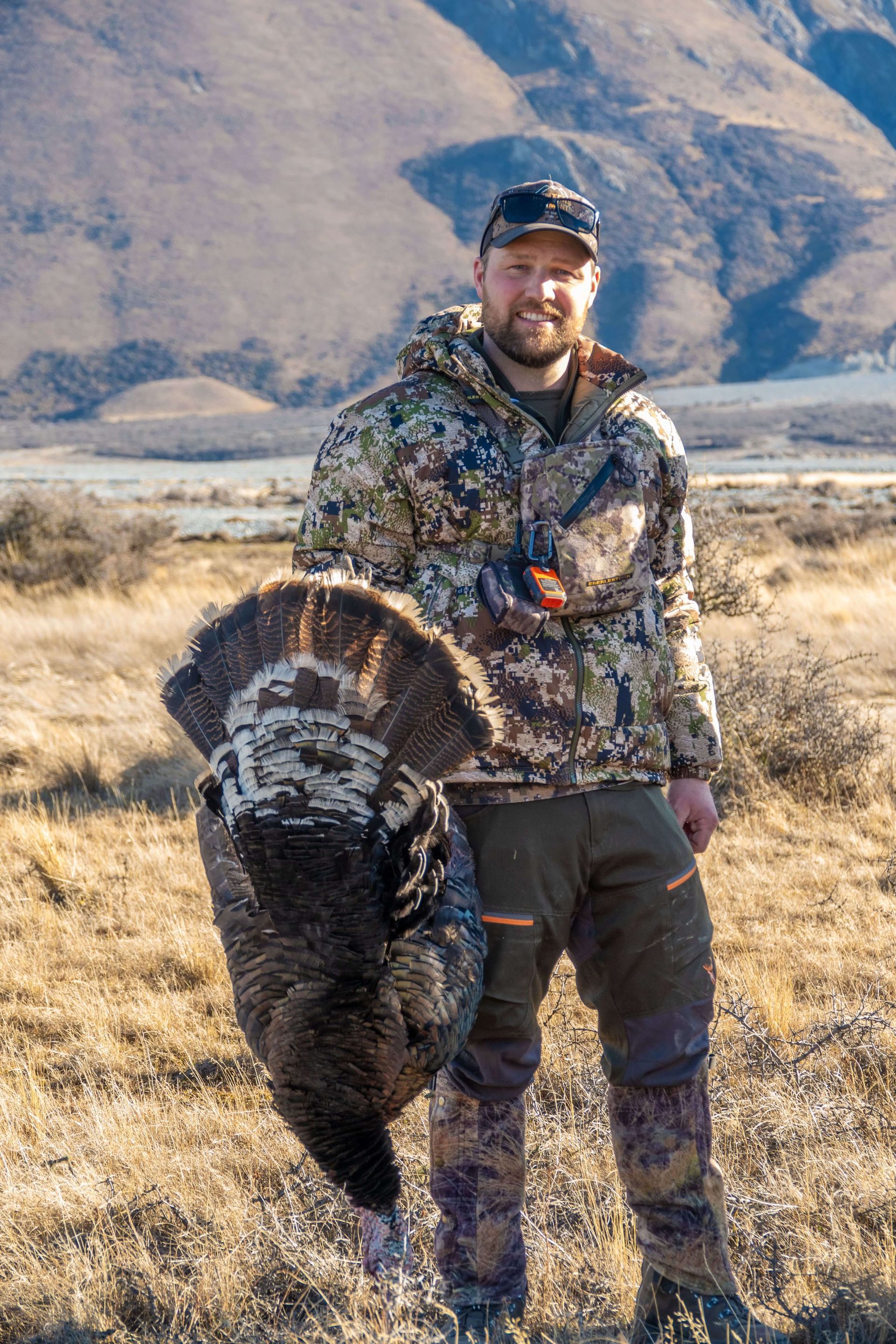 A man in a camouflage jacket is holding a large turkey in a field.