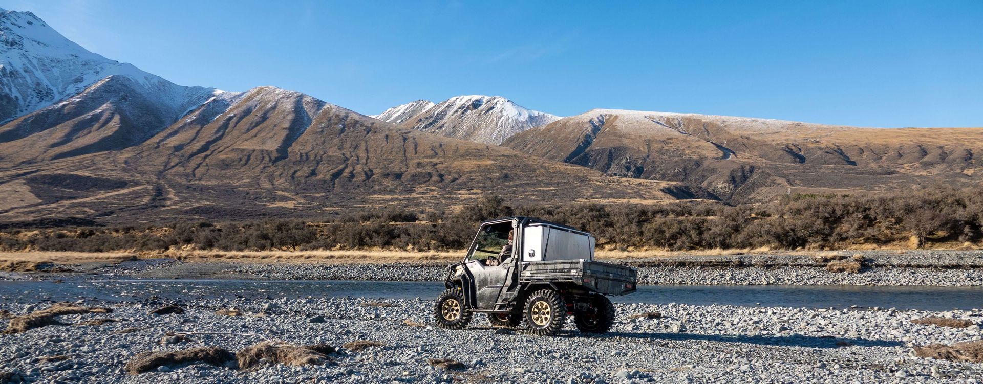 A person is riding a motorcycle on a rocky road next to a river with mountains in the background.