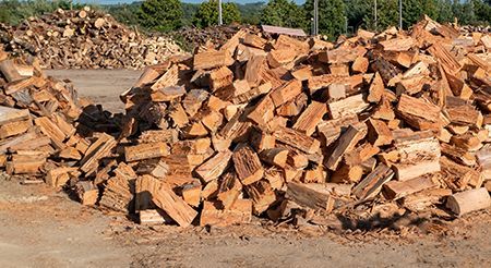 Pile of split firewood outdoors, with additional piles visible in the background.