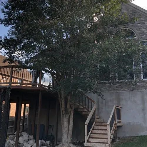 A large tree stands beside a wooden deck and staircase leading to the rear of a multi-story stone house.