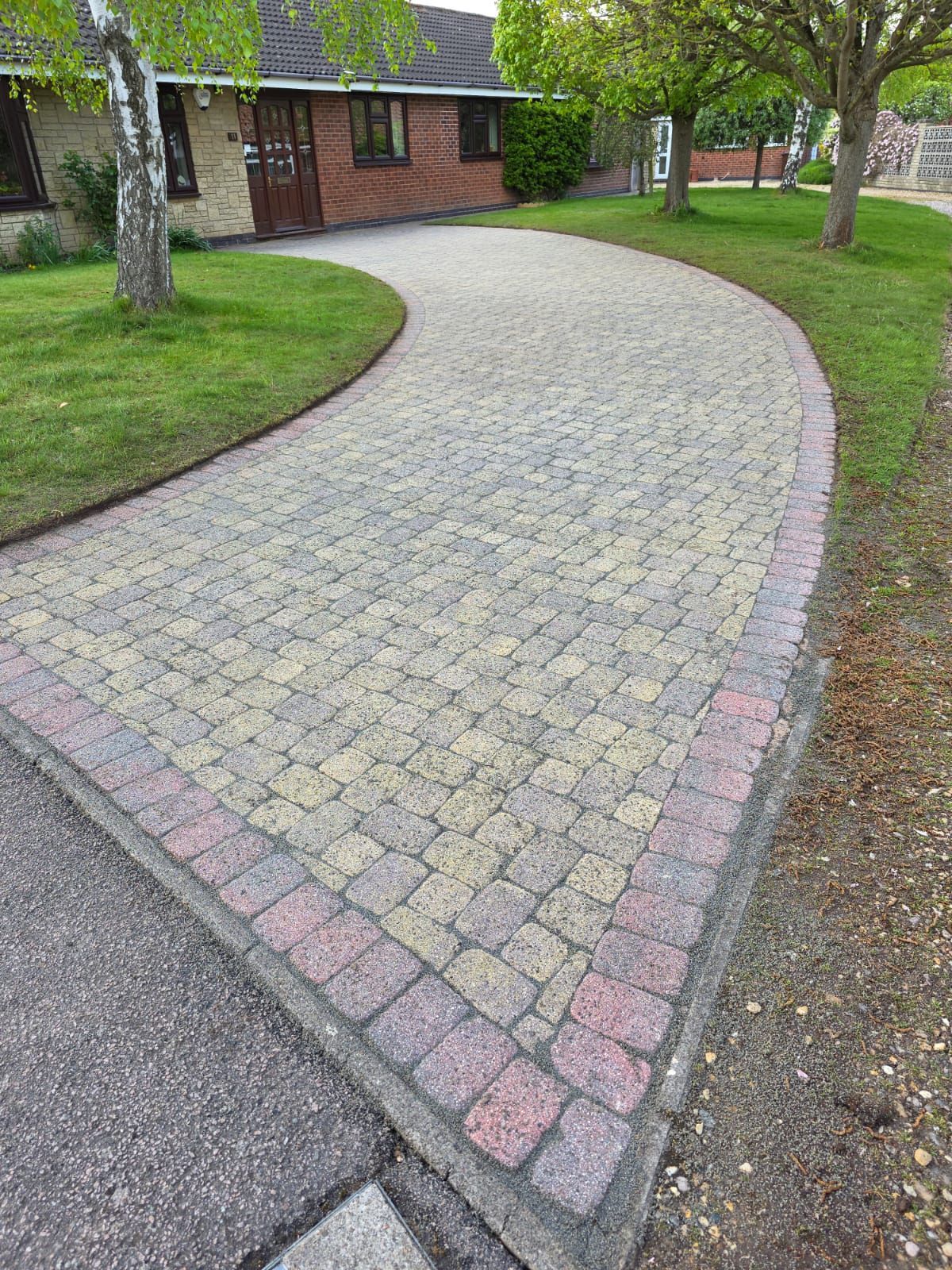 Brick pathway curves towards a house. Green grass borders the bricks, with trees nearby.