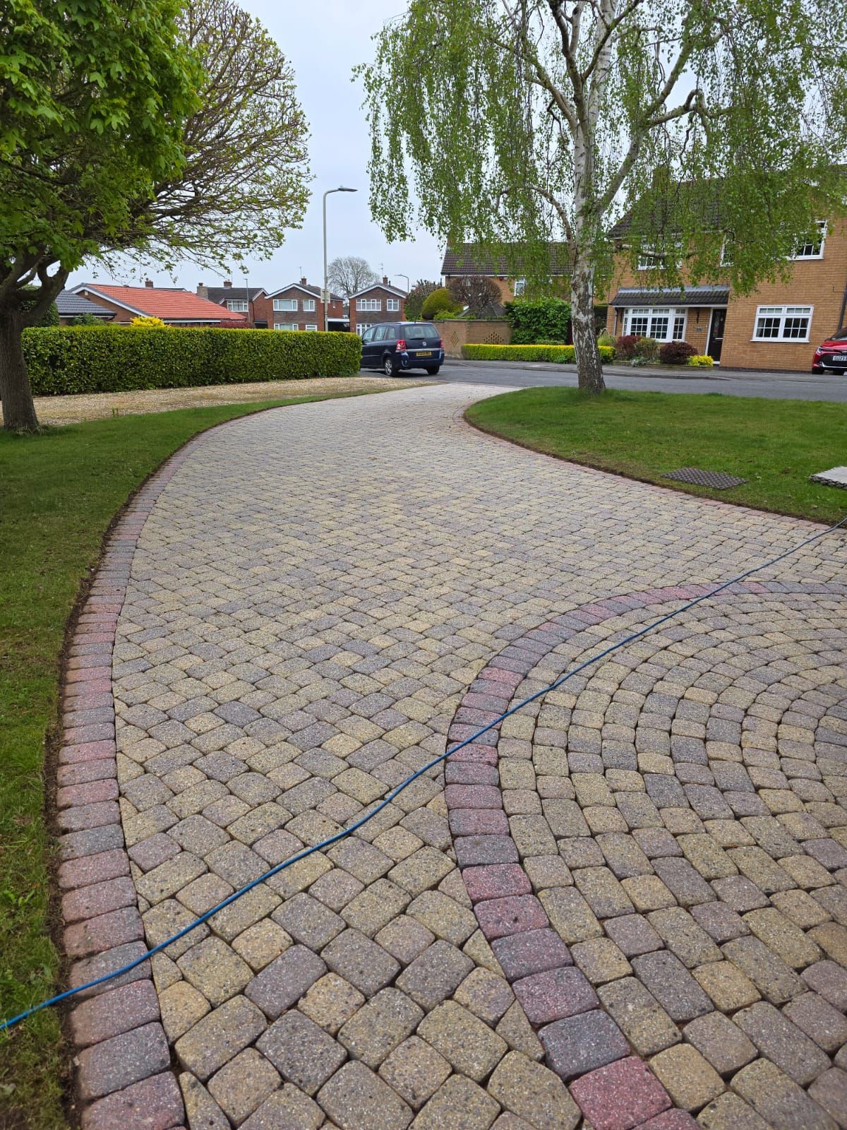 Brick driveway curves through a green lawn. Cars parked in the distance, houses in the background.