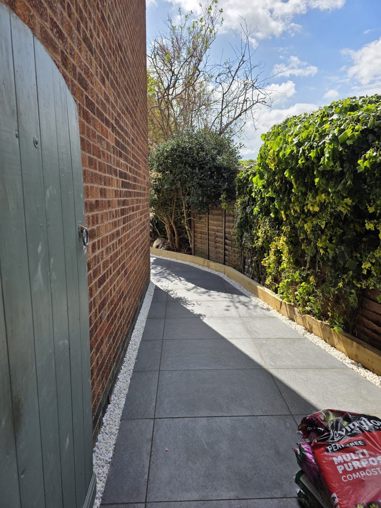 Brick wall, gray paved path, wooden fence, green foliage, blue sky, sunlight.
