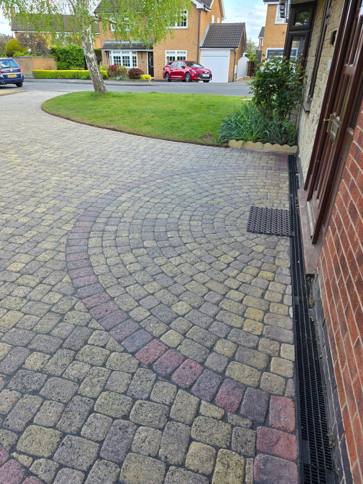 Driveway paved with multi-colored bricks, with a circular patterned section. A house and car are visible in the background.