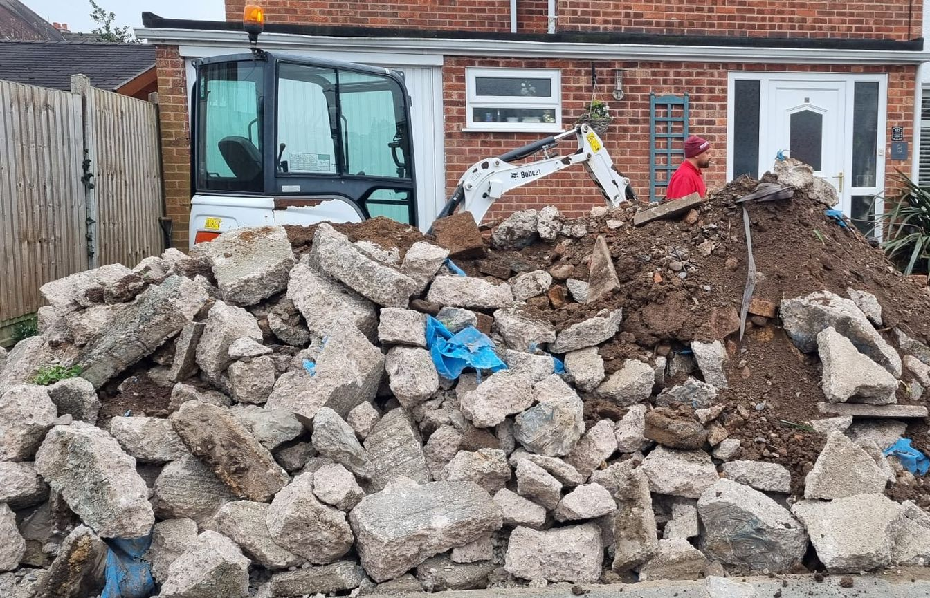 Pile of debris with a white Bobcat excavator in front of a brick house. A person in red is visible.