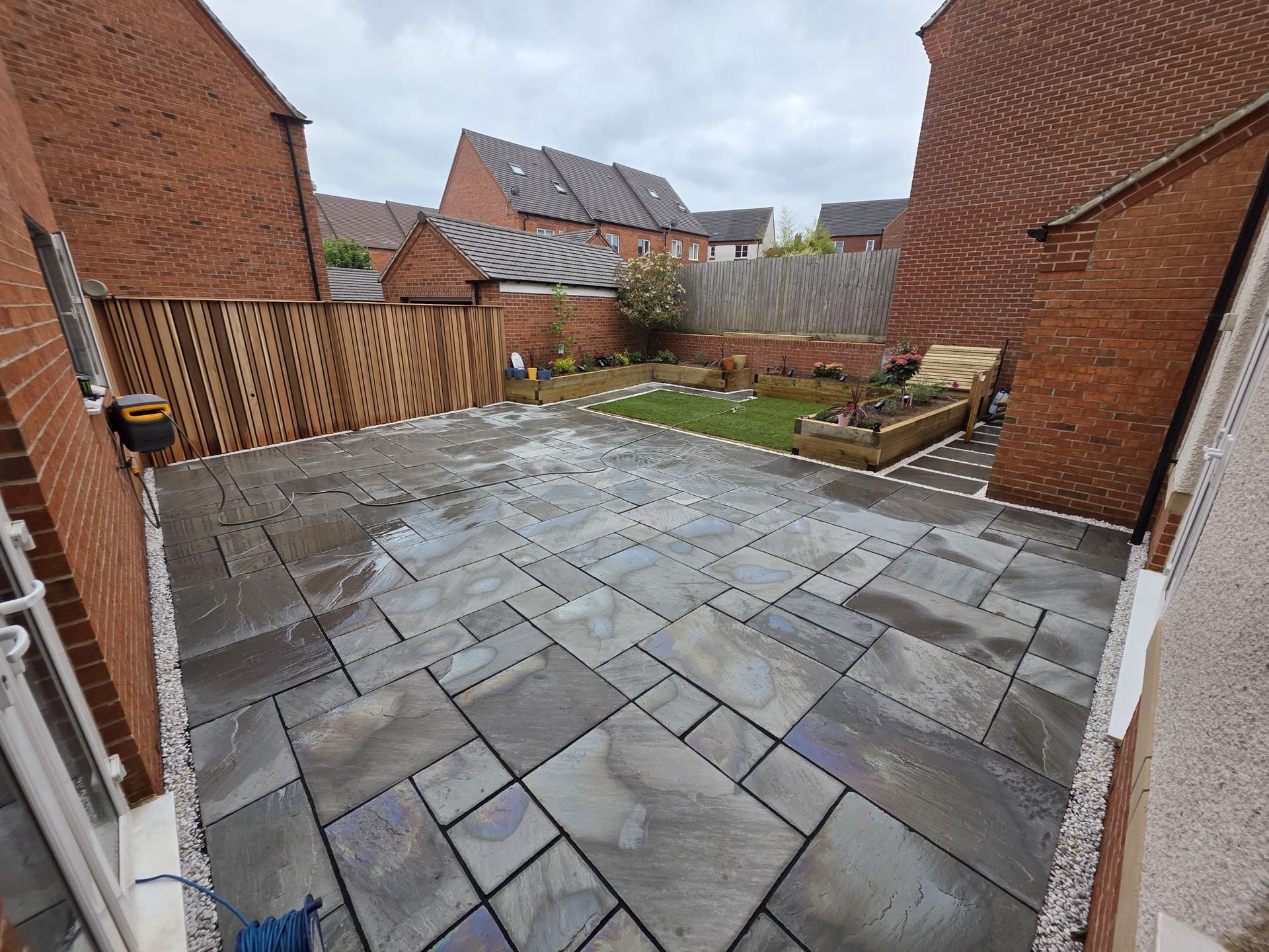 Patio with dark gray paving stones, flanked by brick houses and a wooden fence.