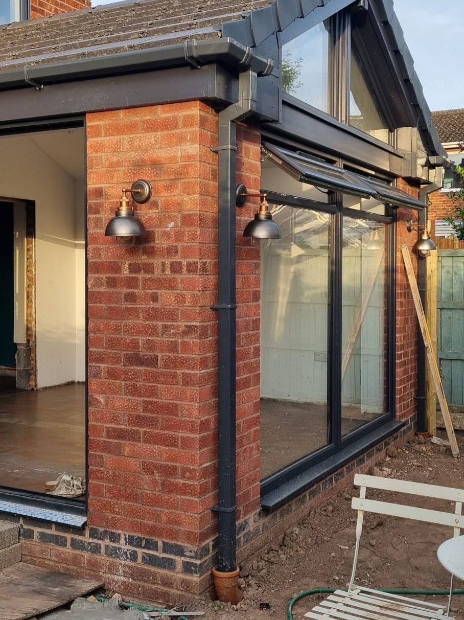 Brick home extension with dark-framed windows, black guttering, and exterior lights.
