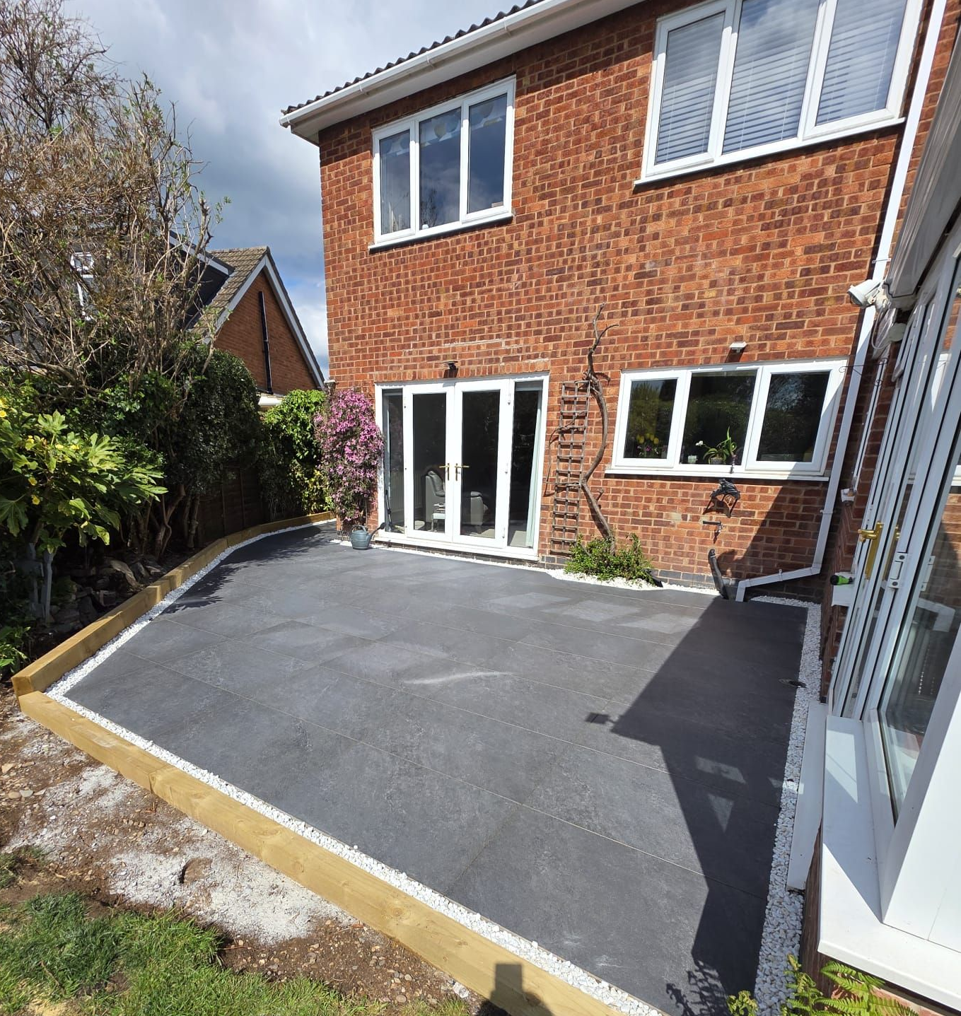 Backyard patio paved with dark grey material, edged with wood and facing a brick house with white-framed windows and doors.