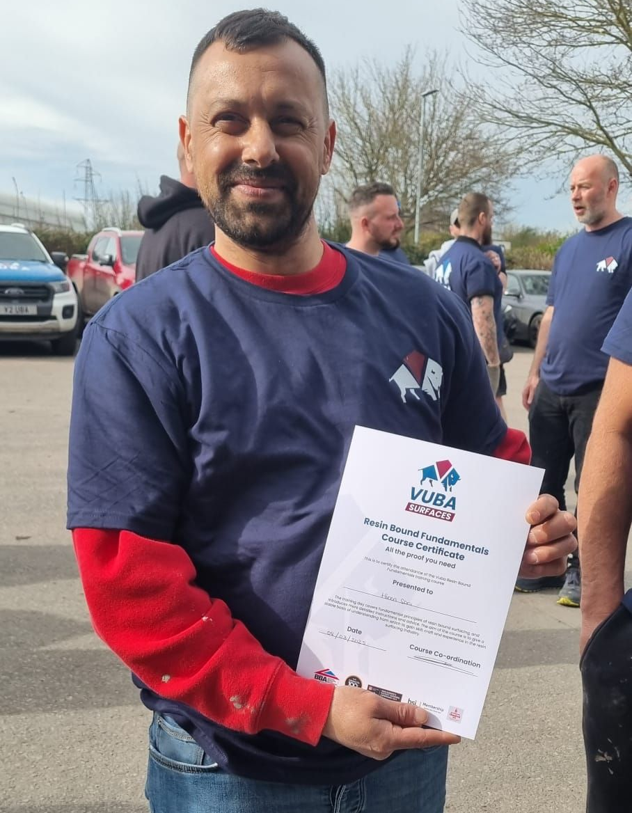 Man in blue shirt, holding certificate, smiling. Others in blue shirts in the background. Outdoors.