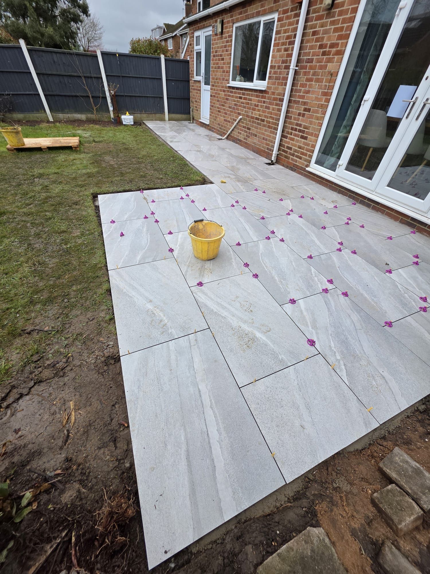 Patio tiles being laid outdoors, with a yellow bucket, on a gray and green setting, near a brick house.