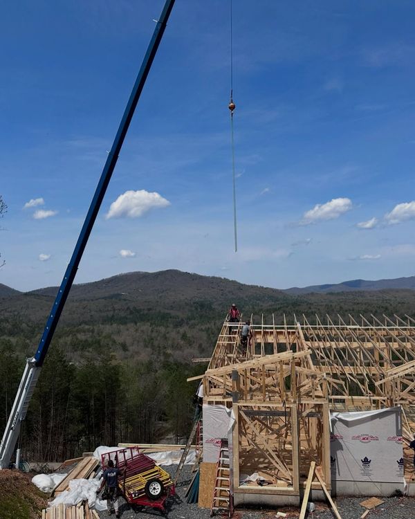 Construction site with crane lifting materials to frame roof, workers on roof, blue sky, mountain view.