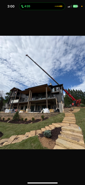 A house under construction with a crane reaching its roof. Stone steps lead up to the house on a grassy hillside.