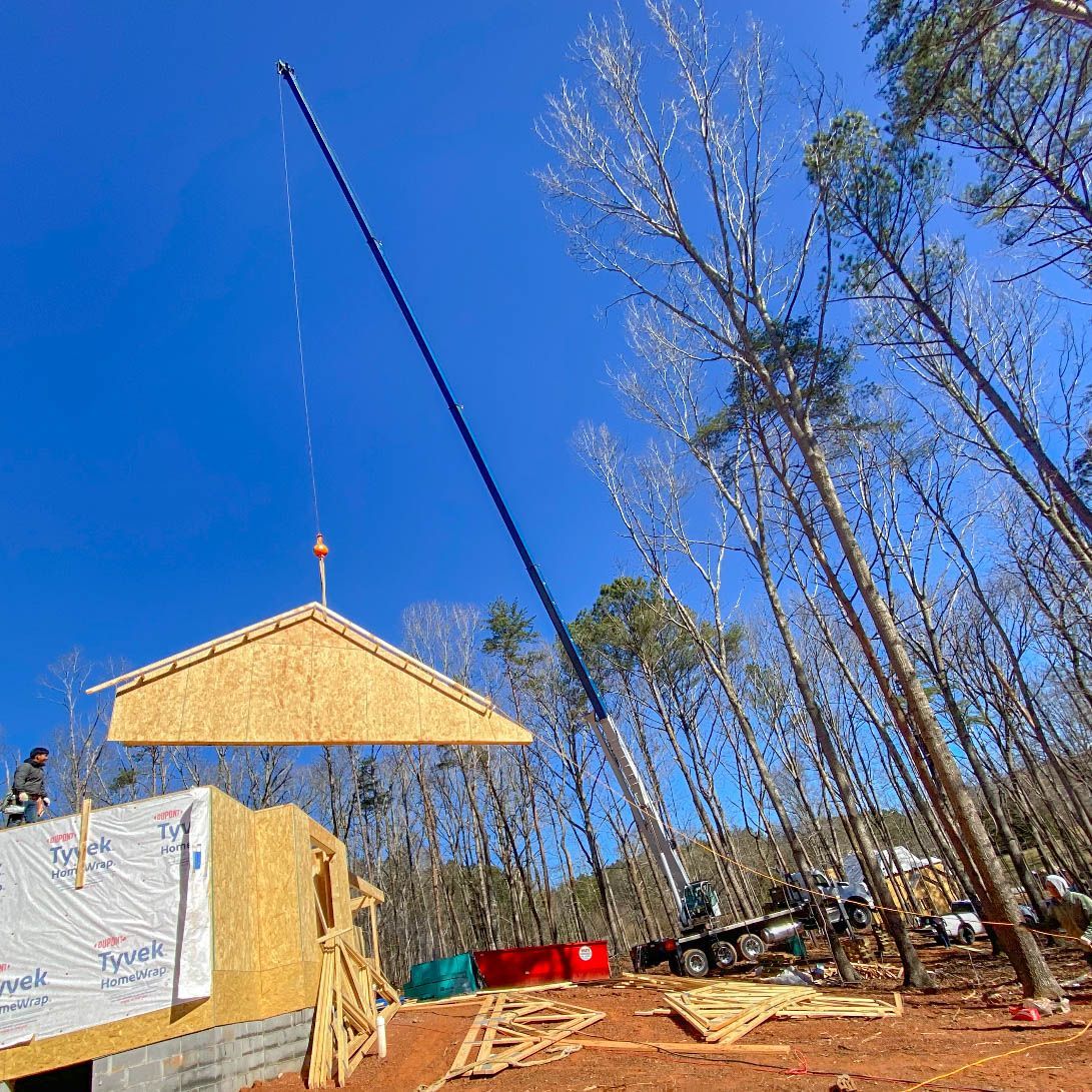 Crane lifting a roof truss onto a wood-framed house under construction on a sunny day.