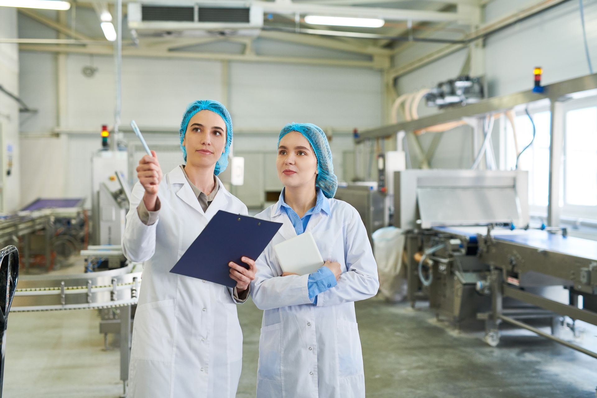 Two women in lab coats and hairnets inspecting a food processing facility. One points upwards, holding a clipboard.