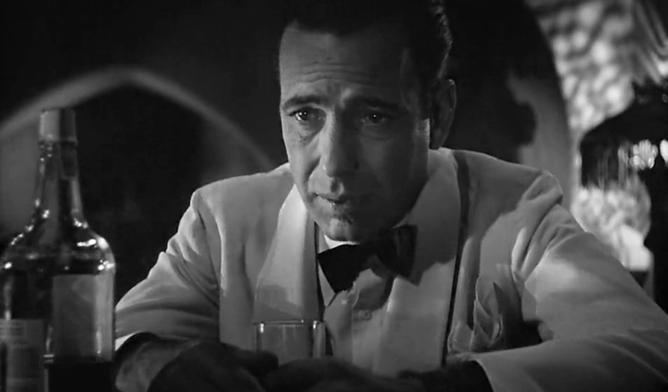 Rick Blaine from Casablanca is sitting at a table with a bottle of alcohol.