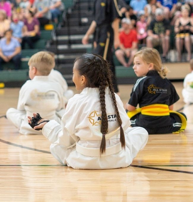 Group of children in martial arts attire posing, some with arms around each other, in a gym.