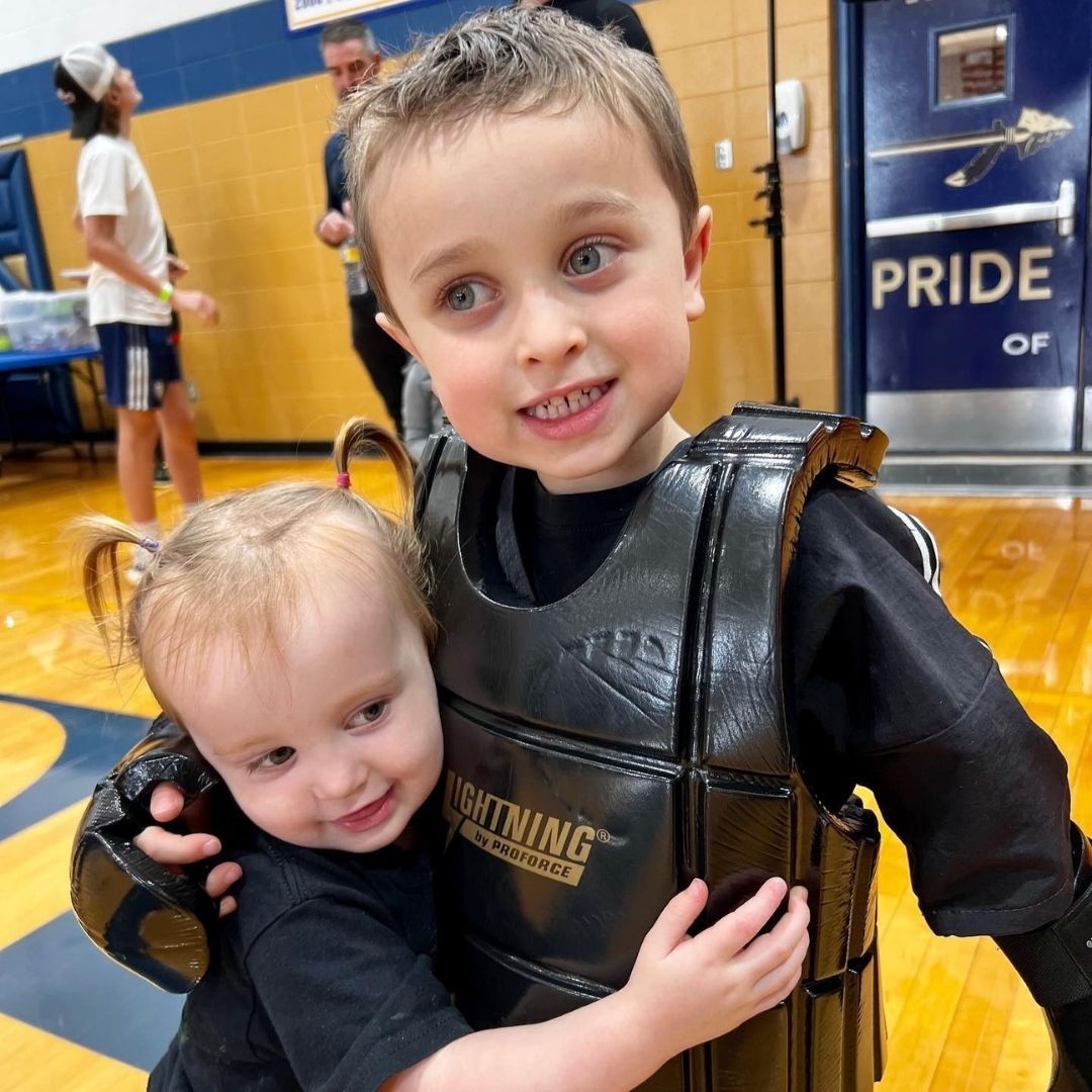 Two men in grappling position on a mat. One embraces the other from behind, both smiling. Gym setting.