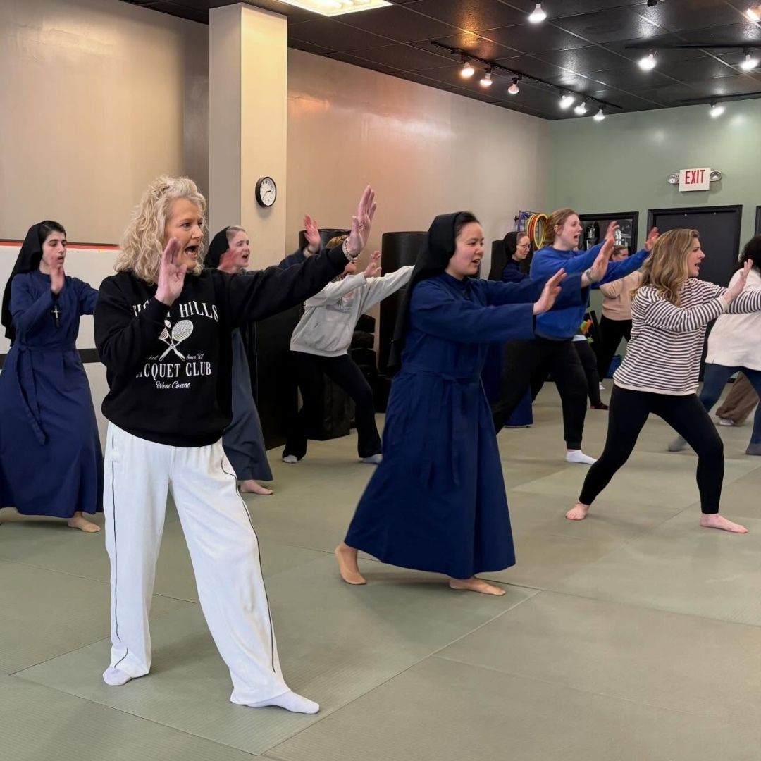 Two children practicing Brazilian Jiu-Jitsu on a mat. One child in white gi has the other in a leg lock. A girl watches.