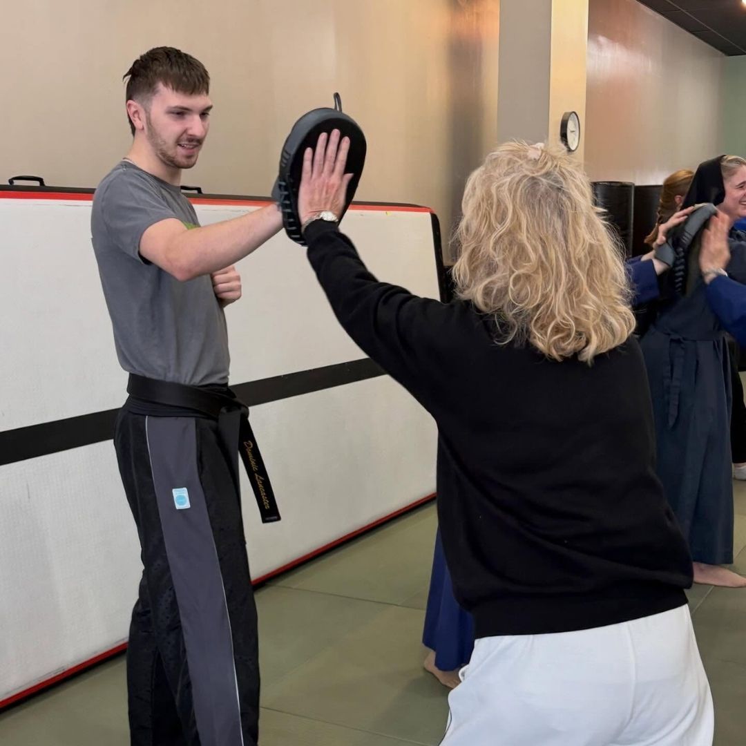 Two young children in blue and black martial arts uniforms grappling on a gym floor, one smiling.