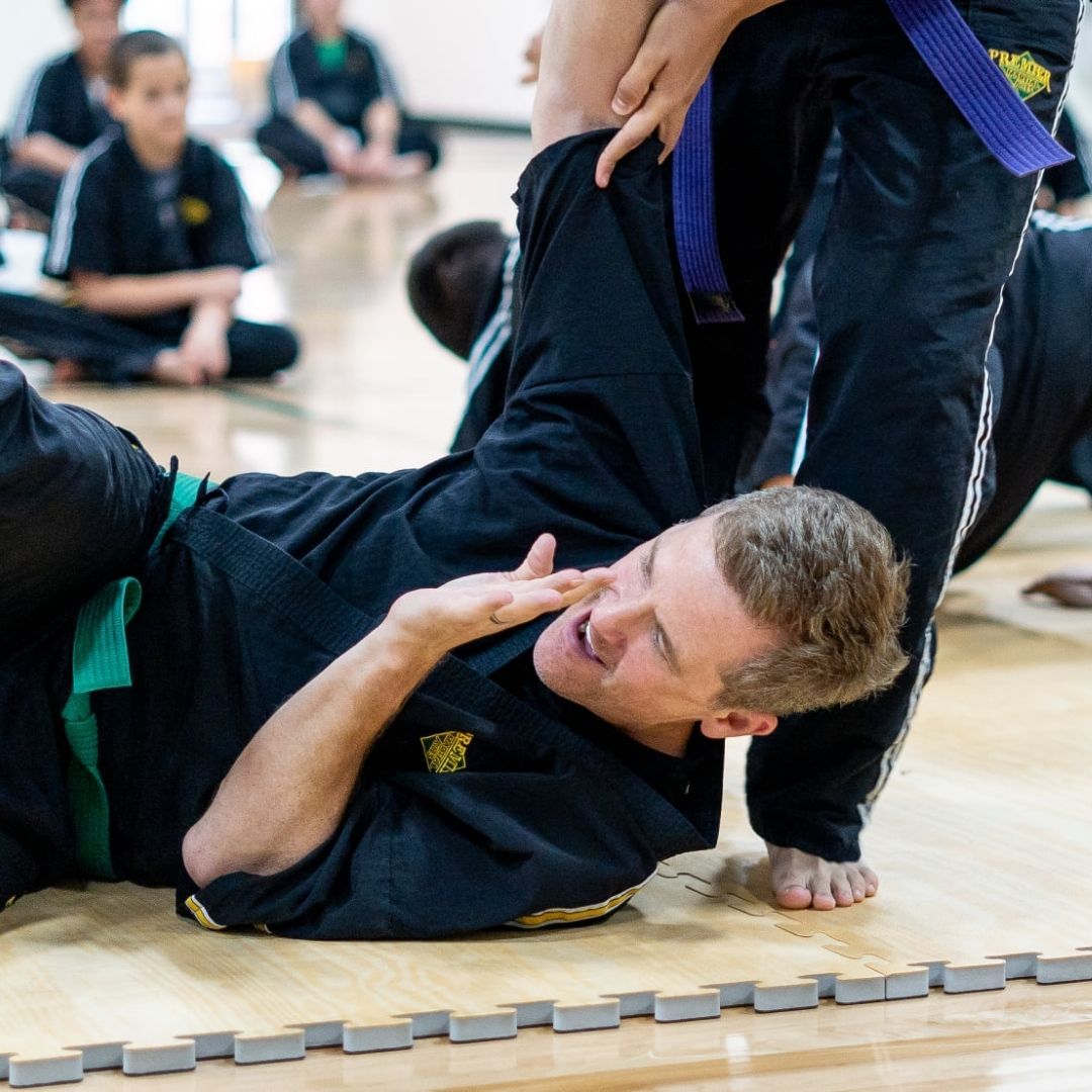 Two children in black and white martial arts uniforms practice a chokehold on a mat. One child smiles.